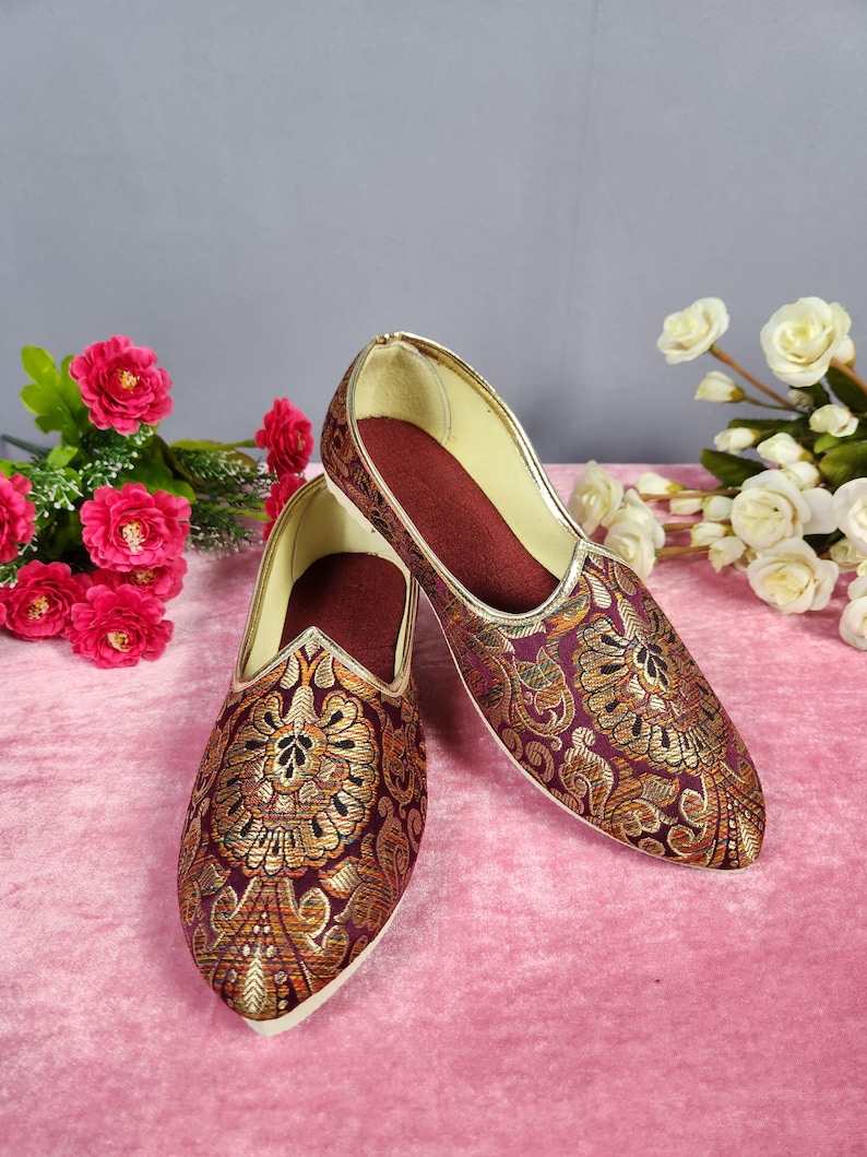 May include: A pair of ornate, slip-on shoes with a pointed toe. The shoes feature a burgundy base with gold floral embroidery. The interior is burgundy, and the trim is gold. The shoes are displayed on a pink surface.