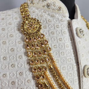 Pu&ograve; includere: Primo piano di una collana dorata con gemme a goccia e fili di perle. La collana &egrave; drappeggiata su una giacca bianca ricamata con bottoni decorativi. Il gioiello presenta un motivo floreale centrale.