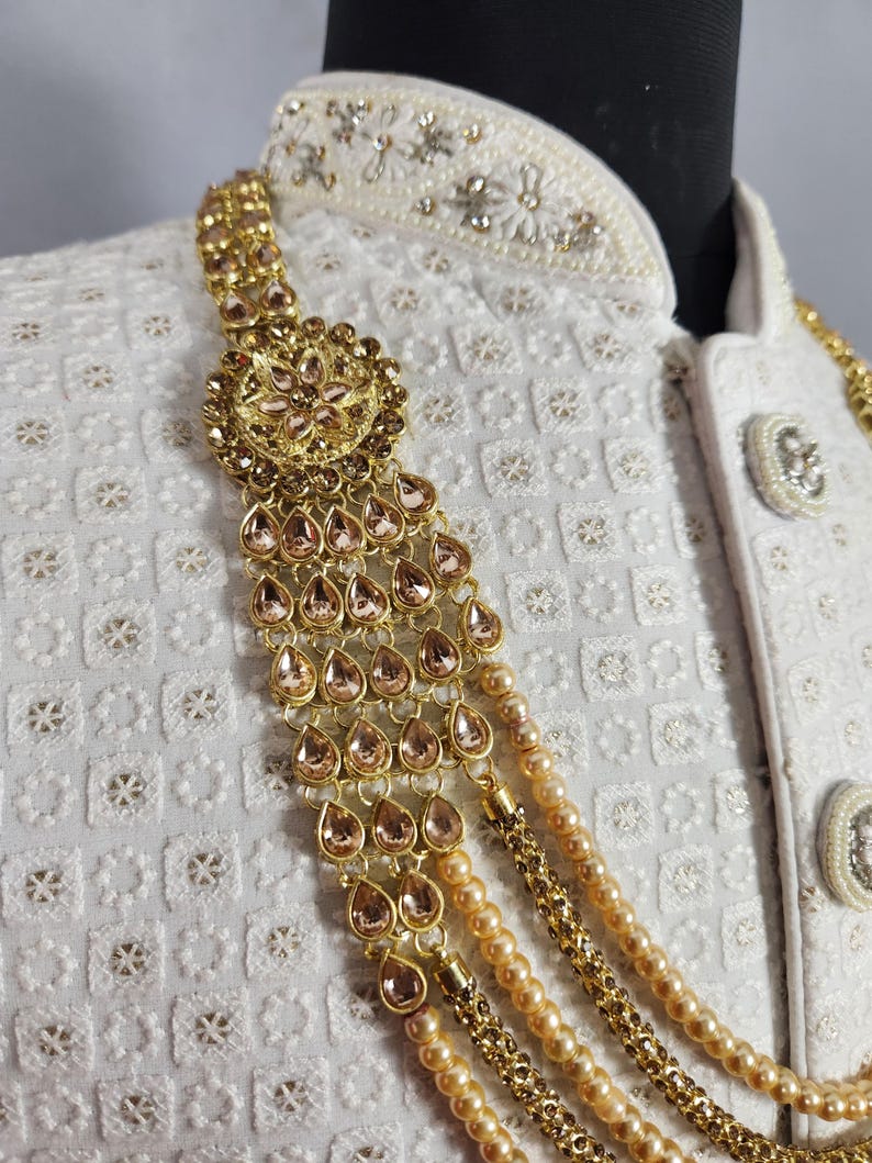 Pu&ograve; includere: Primo piano di una collana dorata con gemme a goccia e fili di perle. La collana &egrave; drappeggiata su una giacca bianca ricamata con bottoni decorativi. Il gioiello presenta un motivo floreale centrale.