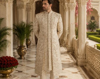 May include: A cream-colored, embroidered sherwani with matching pants and shoes. The long, button-down coat features intricate patterns and is worn by a person standing in a marble hallway with ornate architecture. The outfit is formal and elegant.