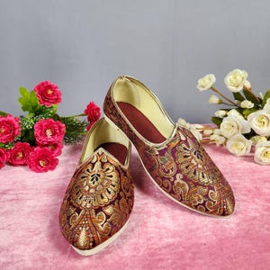 May include: A pair of ornate, slip-on shoes with a burgundy base and gold floral embroidery. The shoes have a pointed toe and a gold trim. The interior is a deep red. The shoes are displayed on a pink surface with floral accents.