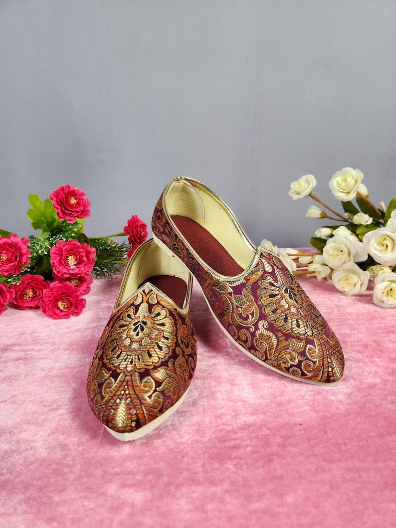 May include: A pair of ornate, slip-on shoes with a burgundy base and gold floral embroidery. The shoes have a pointed toe and a gold trim. The interior is a deep red. The shoes are displayed on a pink surface with floral accents.