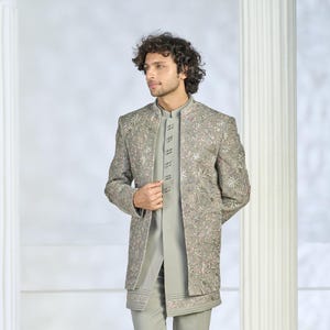 Puede incluir: Chaqueta bordada gris claro con cuello mandar&iacute;n, usada sobre una camisa y pantal&oacute;n gris claro a juego. La chaqueta presenta intrincados bordados florales en rosa y blanco. El atuendo es un conjunto formal.