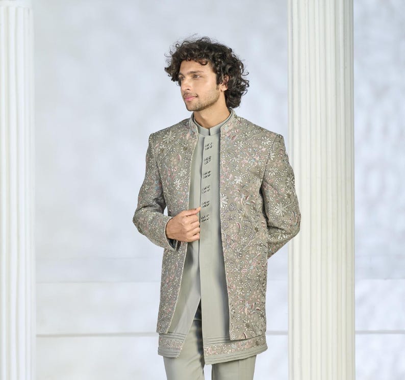 Puede incluir: Chaqueta bordada gris claro con cuello mandar&iacute;n, usada sobre una camisa y pantal&oacute;n gris claro a juego. La chaqueta presenta intrincados bordados florales en rosa y blanco. El atuendo es un conjunto formal.