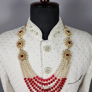 Pu&ograve; includere: Una collana a pi&ugrave; fili con perline rosse e bianche e accenti floreali dorati con pietre rosse. La collana &egrave; esposta su una giacca bianca con ricami intricati.