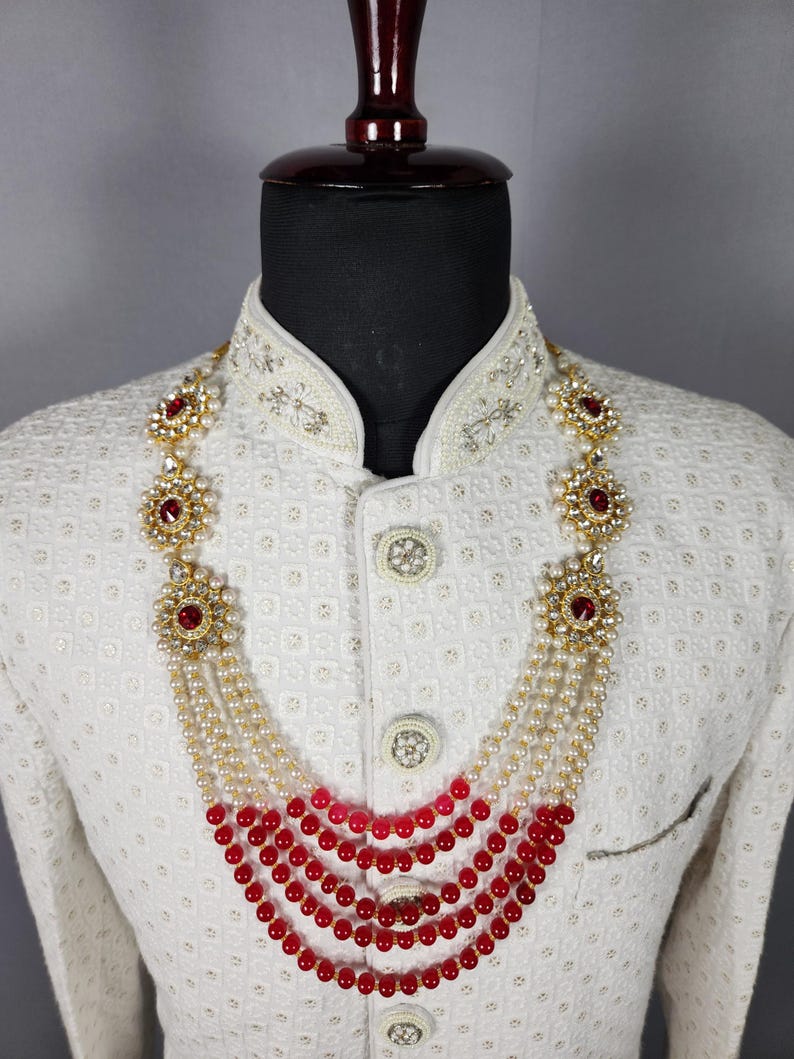 Pu&ograve; includere: Una collana a pi&ugrave; fili con perline rosse e bianche e accenti floreali dorati con pietre rosse. La collana &egrave; esposta su una giacca bianca con ricami intricati.