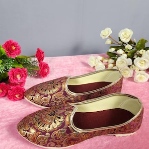 May include: A pair of ornate, slip-on shoes with a burgundy and gold floral pattern. The shoes have a gold trim and a burgundy interior. The background features pink and gray tones, with pink and white flowers.
