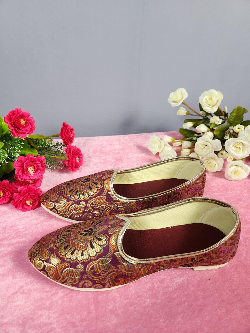 May include: A pair of ornate, slip-on shoes with a burgundy and gold floral pattern. The shoes have a gold trim and a burgundy interior. The background features pink and gray tones, with pink and white flowers.