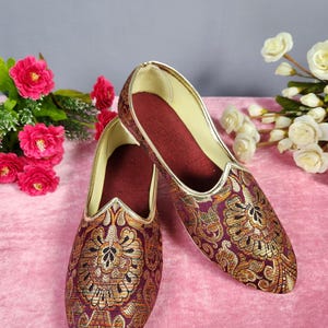 May include: A pair of ornate, slip-on shoes with a pointed toe. The shoes feature a burgundy fabric with gold floral embroidery and a gold trim. The interior is a deep burgundy color. The shoes are likely for formal occasions.