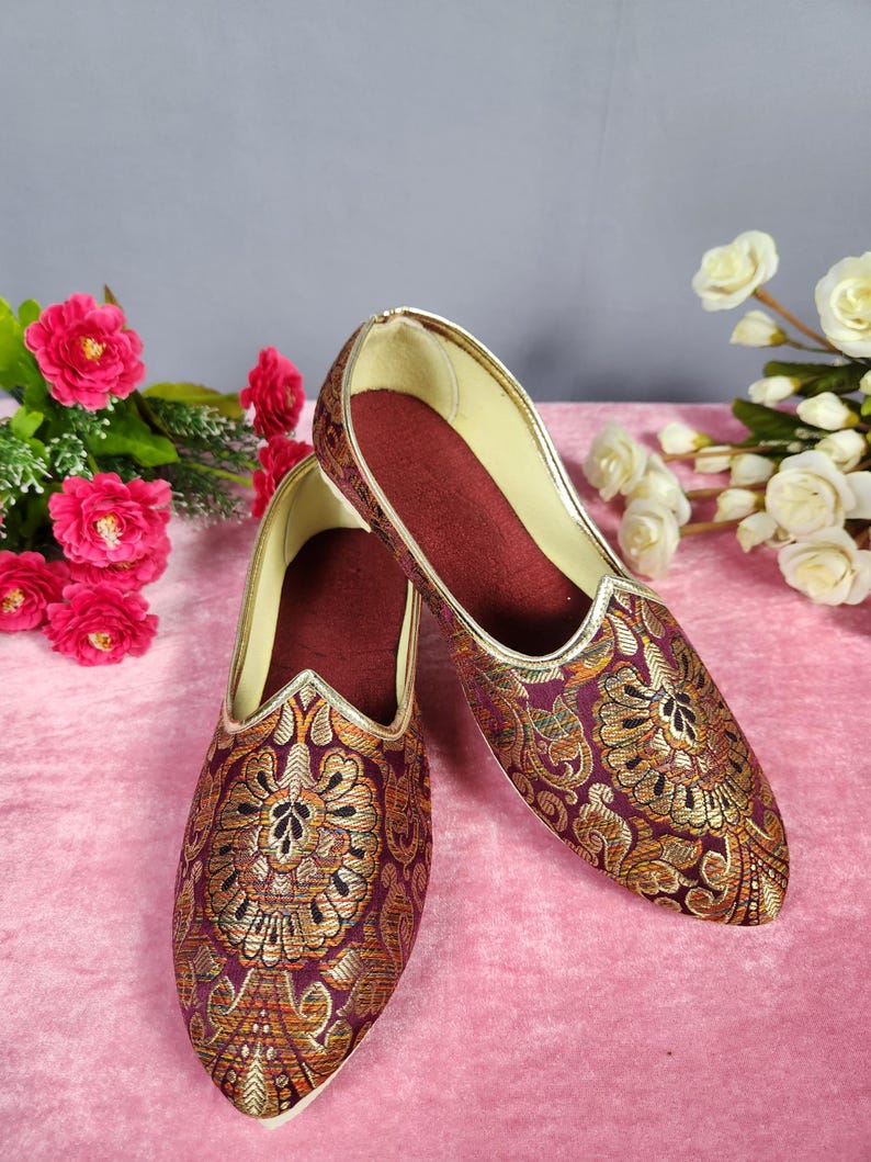 May include: A pair of ornate, slip-on shoes with a pointed toe. The shoes feature a burgundy fabric with gold floral embroidery and a gold trim. The interior is a deep burgundy color. The shoes are likely for formal occasions.