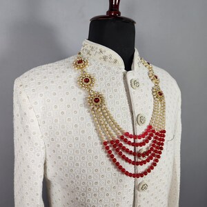 Pu&ograve; includere: Giacca color crema con motivo strutturato adornata da una collana a pi&ugrave; fili. La collana presenta perline rosse, perle e dettagli dorati. La giacca ha bottoni decorativi e un colletto alto.