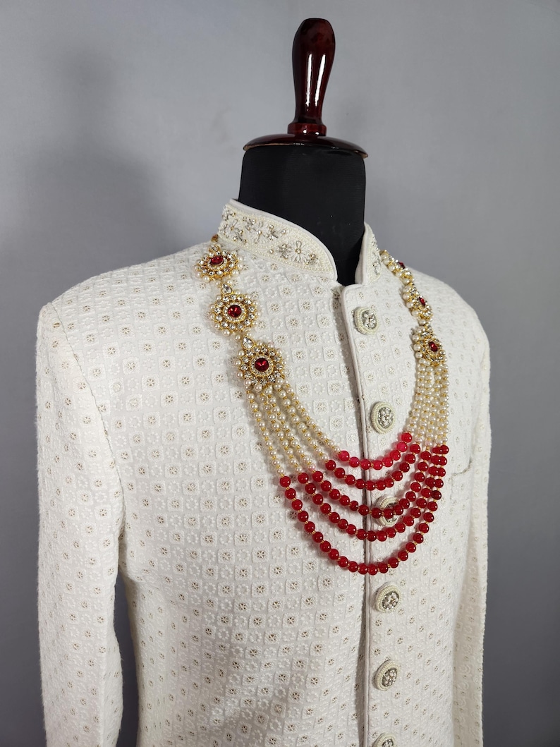 Pu&ograve; includere: Giacca color crema con motivo strutturato adornata da una collana a pi&ugrave; fili. La collana presenta perline rosse, perle e dettagli dorati. La giacca ha bottoni decorativi e un colletto alto.