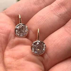 May include: A pair of gold leverback earrings, each featuring a single round, faceted clear stone set in a prong setting. The earrings have a polished gold finish and a classic circular design, showcasing a bright, reflective surface on the stones.