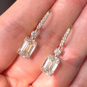 May include: A pair of drop earrings crafted in yellow gold, each featuring a large emerald-cut center stone suspended from a smaller round brilliant-cut diamond, with a slender, vertical bar set with a row of tiny round diamonds extending upward.