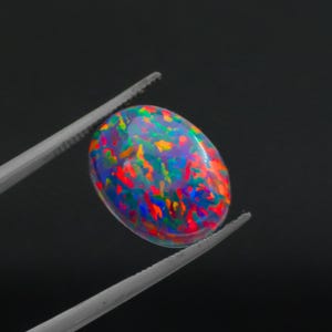 Könnte beinhalten: Ein ovaler Opal-Edelstein mit einer lebendigen Farbpalette, darunter Rot, Orange, Gelb, Grün und Blau. Der Edelstein wird von einer silbernen Pinzette vor schwarzem Hintergrund gehalten.