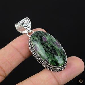 May include: Oval pendant with a green and black gemstone with red inclusions, set in a silver-toned metal frame. The pendant has a decorative bail and a detailed border. The gemstone is held in a hand.