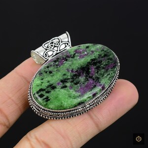 Könnte beinhalten: Ovaler Anhänger mit einem grün-violett gesprenkelten Stein, gefasst in silberfarbenem Metall. Das Muster des Steins umfasst Grün, Lila und Schwarz. Der Anhänger hat eine dekorative Öse oben für eine Kette.