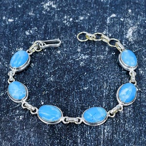 Könnte beinhalten: Silberarmband mit ovalen, hellblauen Edelsteinen. Die Edelsteine sind in einen silbernen Metallrahmen gefasst und durch silberne Glieder verbunden. Das Armband hat einen Verschluss. Auf dunkler, strukturierter Oberfläche.
