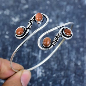 Peut inclure: Bracelet manchette argenté avec trois pierres précieuses ovales brun-rouge. Le bracelet présente un design ouvert unique avec des volutes décoratives et un motif texturé près des pierres précieuses. Le bracelet est tenu par une main.