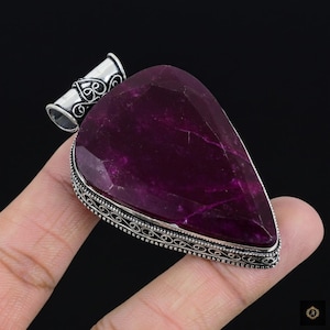 May include: A large, teardrop-shaped pendant with a faceted, deep magenta gemstone. The stone is set in a silver-colored metal with an ornate, filigree border. The bail is also silver-colored with decorative scrollwork.