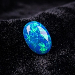 May include: An oval-shaped cabochon gemstone featuring a deep blue base color with vibrant, iridescent flashes of bright green and light blue light throughout the surface, displaying a high-gloss finish with visible light reflections.