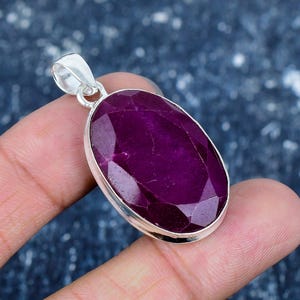Op de afbeelding: Zilveren hanger met een grote, facetgeslepen, ovaalvormige, diep magenta edelsteen. De edelsteen is gezet in een gepolijste zilveren bezel met een zilveren bale. De hanger wordt in een hand gehouden.