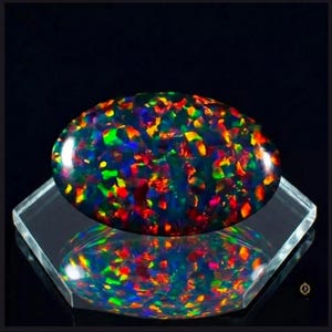 Könnte beinhalten: Ein ovaler schwarzer Opal-Edelstein mit einem lebendigen Farbspiel aus verstreuten roten, orangen, gelben, grünen und blauen Lichtreflexen. Der polierte Stein liegt auf einem klaren, sechseckigen Acryl-Präsentationsständer und spiegelt sich.