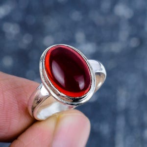 May include: A silver ring with a large, oval, deep red gemstone. The gemstone is set in a bezel setting, and the ring has a simple, polished band. The ring is held by a person's fingers.