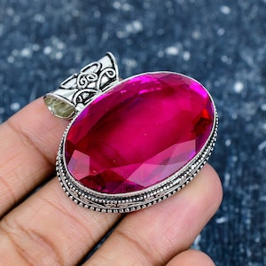 Könnte beinhalten: Ein ovaler Anhänger mit einem facettierten, magentafarbenen Edelstein. Der Stein ist in silberfarbenem Metall mit detaillierten Filigranarbeiten an den Rändern und einem dekorativen Bügel gefasst. Ca. 3 cm lang.