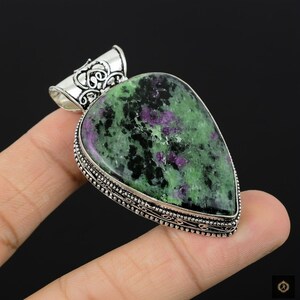 Könnte beinhalten: Ein großer, tropfenförmiger Anhänger mit einer silbernen Öse. Der Anhänger zeigt einen polierten Edelstein mit einer Mischung aus grünen, schwarzen und violetten Farbtönen. Der Edelstein ist in einen dekorativen Silberrahmen gefasst.