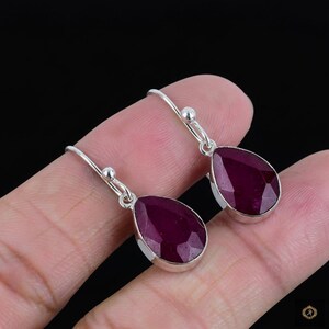 May include: A pair of silver drop earrings featuring faceted, teardrop-shaped, deep red gemstones. The earrings have a simple, elegant design with a small ball detail at the top of the hook.