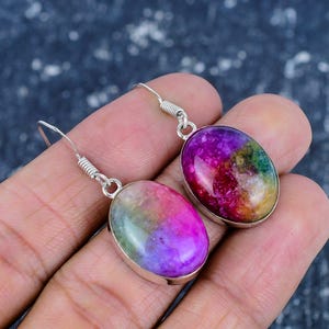 Könnte beinhalten: Ein Paar ovale Ohrringe mit einem lebendigen, mehrfarbigen Stein. Die Steine zeigen Farbtöne von Rosa, Lila, Grün und Gelb, gefasst in einem silberfarbenen Metall. Die Ohrringe haben einen Hakenverschluss.