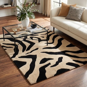 Pu&ograve; includere: Un tappeto con stampa zebrata beige e nera. Il tappeto rettangolare presenta audaci strisce nere su uno sfondo neutro. &Egrave; in un soggiorno, con un tavolino con piano in vetro e un divano bianco. Il design crea un contrasto sorprendente.
