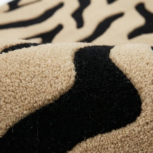 Pu&ograve; includere: Primo piano di un tappeto beige e nero a righe zebrate. Il tappeto ha una superficie morbida e strutturata con fibre spesse e soffici. Le strisce nere sono audaci e irregolari, in contrasto con lo sfondo beige pi&ugrave; chiaro. Il tappeto sembra essere piegato.