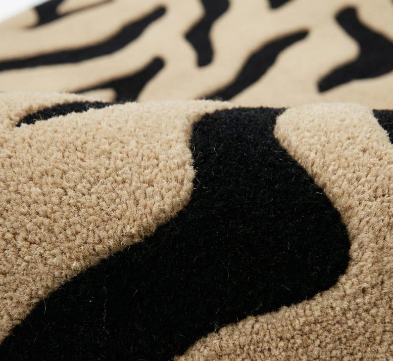 Pu&ograve; includere: Primo piano di un tappeto beige e nero a righe zebrate. Il tappeto ha una superficie morbida e strutturata con fibre spesse e soffici. Le strisce nere sono audaci e irregolari, in contrasto con lo sfondo beige pi&ugrave; chiaro. Il tappeto sembra essere piegato.