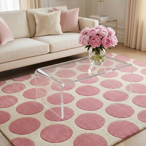 Könnte beinhalten: Ein Wohnzimmer mit cremefarbenem Sofa, rosa Kissen und einem transparenten Couchtisch aus Acryl. Der Teppich hat einen cremefarbenen Hintergrund mit großen rosa Punkten. Eine Glasvase enthält rosa Pfingstrosen.