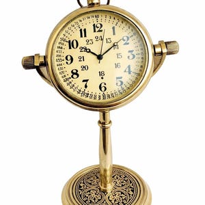 Puede incluir: Un reloj de escritorio de estilo vintage, de color dorado. El reloj tiene una esfera redonda con números y manecillas negras, y un anillo interior más pequeño con números adicionales. Se apoya sobre una base decorativa.