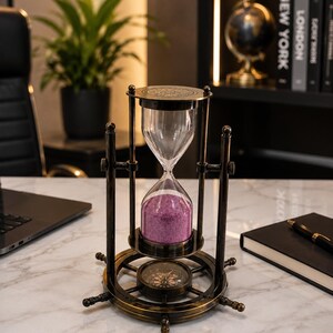 May include: A vintage-style hourglass featuring a dark brass frame with a ship's wheel base containing an integrated compass. The glass bulb is filled with fine purple sand, held between two circular brass plates with decorative engravings.