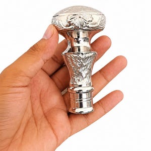May include: A polished silver-colored metal walking stick handle featuring an ornate, embossed floral pattern on the rounded top and a tapered, textured shaft. The handle has a smooth, reflective finish and a cylindrical base with horizontal ridges.