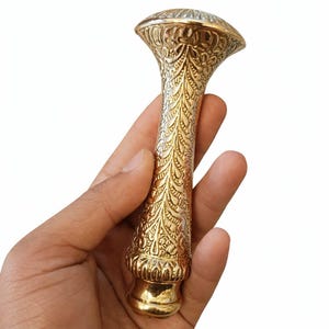 May include: Ornate gold-colored metal cane handle with detailed floral and leaf patterns. The handle has a bulbous top and a cylindrical shaft, held in a hand against a white background. The design is intricate and decorative.