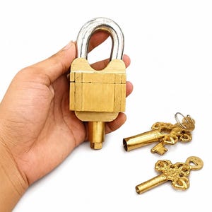 Peut inclure: Un cadenas en laiton avec une anse en métal argenté et une saillie cylindrique à la base, accompagné de deux clés en laiton ornées dotées de têtes en filigrane complexe et de tiges creuses, le tout sur un fond blanc uni.