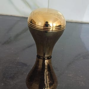 Peut inclure: Une poignée en laiton poli avec un sommet arrondi en forme de dôme et un corps effilé en forme de sablier. La surface métallique est très réfléchissante, présentant une finition dorée avec des lignes gravées horizontales près des extrémités.