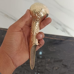 May include: A gold-coloured finger guard with an ornate floral design. The finger guard has a rounded top and a long, pointed blade. The metal has a polished finish, and the design is engraved into the surface.