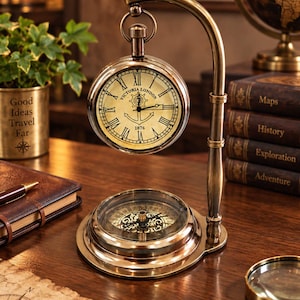 May include: A brass-toned desk clock and compass set featuring a hanging pocket watch style clock with Roman numerals and the text Victoria London 1876, suspended from a curved stand above a circular magnetic compass base with a glass cover.