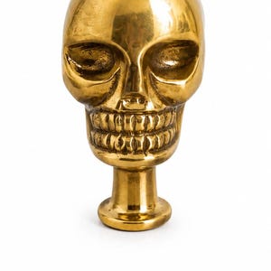 Peut inclure: Une sculpture de crâne en métal doré poli présentant un crâne lisse et arrondi, des orbites profondes et une mâchoire détaillée avec des dents visibles. Le crâne est monté sur une courte base cylindrique avec un fond circulaire plat.