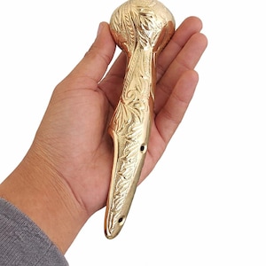 May include: A gold-colored, ornate handle with a rounded top and a curved, elongated base. The surface is covered in detailed floral and leaf patterns. The handle has two small holes near the base.
