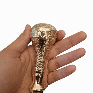Op de afbeelding: Een messing wandelstokhandvat met ingewikkelde bloemen- en geometrische patronen. De handgreep heeft een afgeronde bovenkant met gegraveerde details en een gepolijste afwerking. De handgreep wordt in een hand gehouden tegen een witte achtergrond.