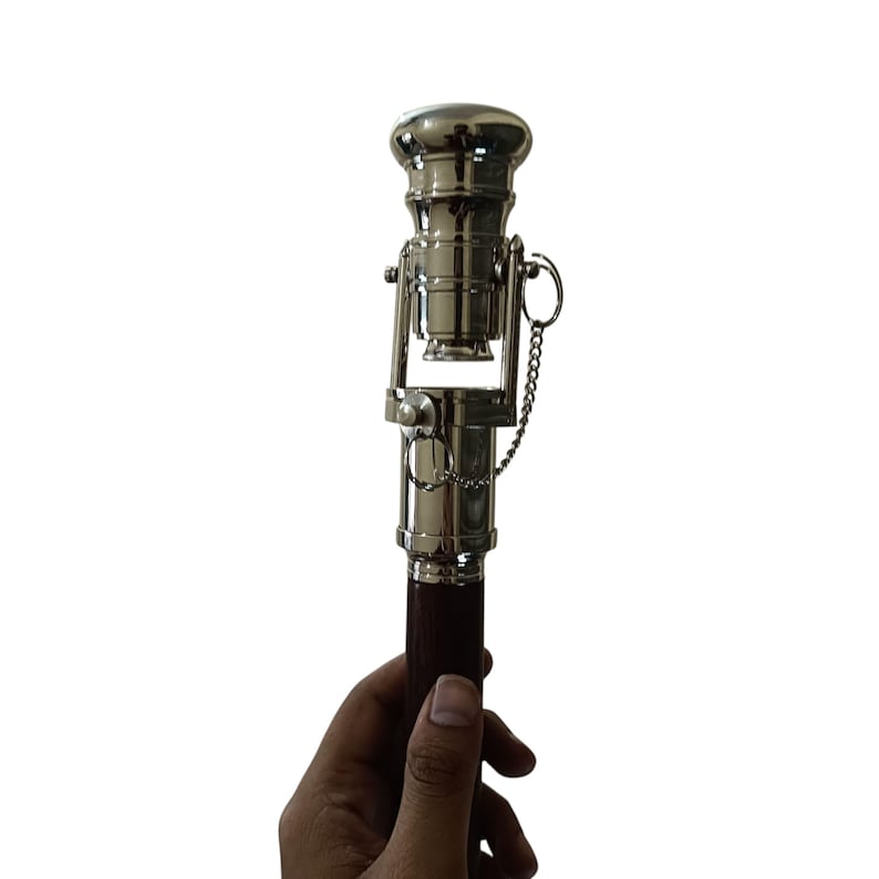 Pu&ograve; includere: Un bastone da passeggio in stile vintage color argento con un manico in legno marrone scuro. La sezione superiore ha un design a forma di telescopio con una piccola catena e un dettaglio ad anello. Il bastone &egrave; tenuto in mano su sfondo bianco.
