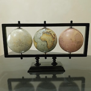 May include: A decorative globe set featuring three globes in a black frame. The globes are in cream, blue-green, and pink, each with a vintage map design. The base and frame are black, creating a striking contrast.