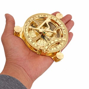 May include: A polished gold-tone brass sundial compass featuring Roman numerals, an adjustable gnomon, and engraved text reading "W.W. London". The circular instrument includes two side adjustment knobs and a central magnetic needle.